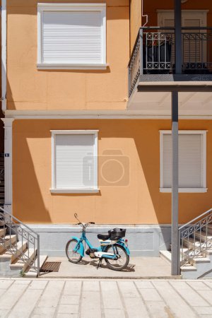 A retro blue moped stands parked on a sunny street against a vibrant yellow building with white windows and balcony.