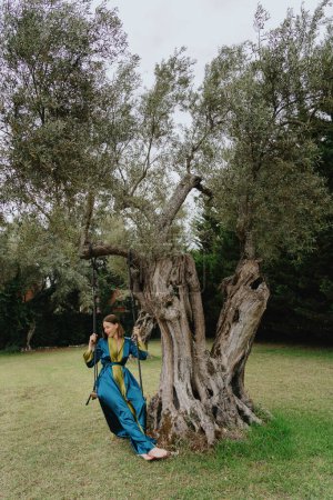 A joyful woman in a flowing blue dress swings from an ancient olive tree in a lush green field under the clear sky.