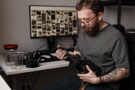 Foto de Fotógrafo masculino enfocado limpiando cuidadosamente la lente de una cámara profesional en su espacio de trabajo organizado y contemporáneo. - Imagen libre de derechos