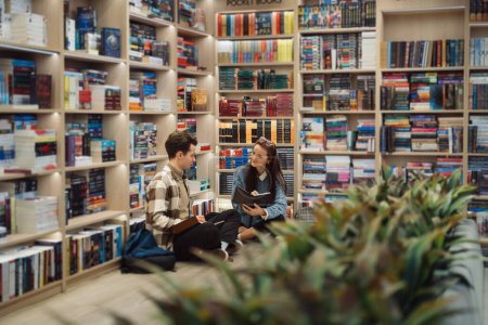 A young man and woman sit on the floor of a well-stocked library, engaged in a discussion over a book. They are surrounded by shelves filled with a variety of books.