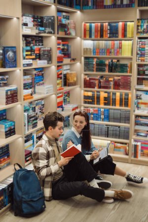 A young man and woman sit on the floor of a well-stocked bookstore, absorbed in reading books. A casual, intellectual outing highlighting love for literature.