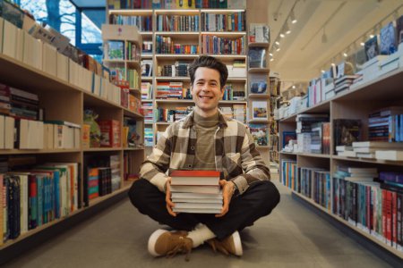A cheerful young man sits on the floor of a well-lit bookstore, surrounded by shelves packed with diverse titles. He holds a stack of books, smiling contentedly.
