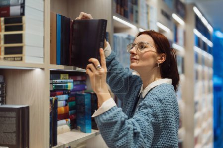 Photo for Focused young woman with glasses selects a book from a shelf in a well-lit library. Her interest and curiosity are evident as she explores different titles. - Royalty Free Image