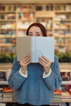 Eine junge rothaarige Frau bedeckt ihr Gesicht mit einem Buch in einer gemütlichen Bibliotheksatmosphäre, umgeben von Regalen voller Bücher.