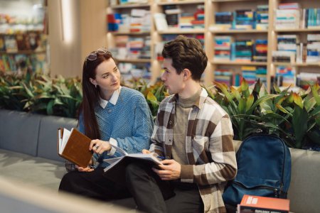 A young man and woman sit comfortably in a modern bookshop, surrounded by books, deeply engaged in a conversational exchange. Their expression shows interest and thoughtfulness.