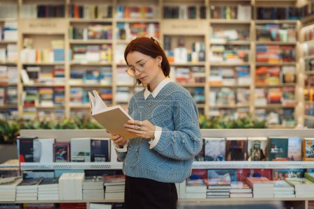 Une jeune femme concentrée dans des lunettes et un pull bleu lit un livre tout en se tenant dans une librairie dynamique et bien approvisionnée. Les étagères sont remplies d'une variété de livres.