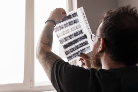 Primer plano de un fotógrafo barbudo sosteniendo e inspeccionando negativos de película en luz natural por una ventana.