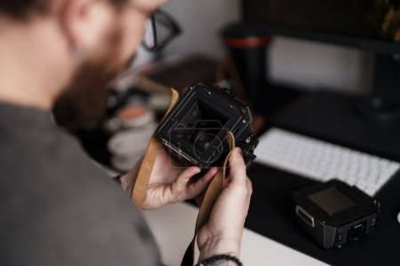 Foto de Primer plano de un fotógrafo profesional inspeccionando cuidadosamente los detalles de una cámara de formato medio con un telón de fondo de oficina moderno. - Imagen libre de derechos