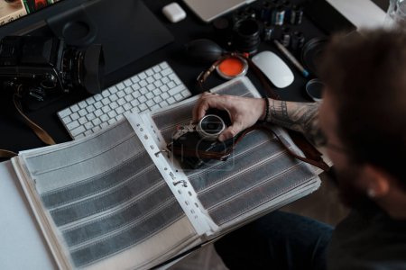 Ein tätowierter Fotograf untersucht Filmnegative sorgfältig an einem gut organisierten Schreibtisch mit Fotoausrüstung..