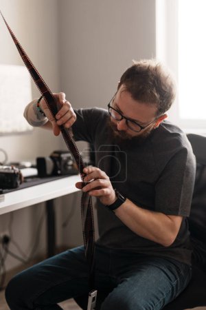 Fotógrafo masculino enfocado que examina negativos de película en un espacio de trabajo moderno y bien iluminado con una cámara en el escritorio.