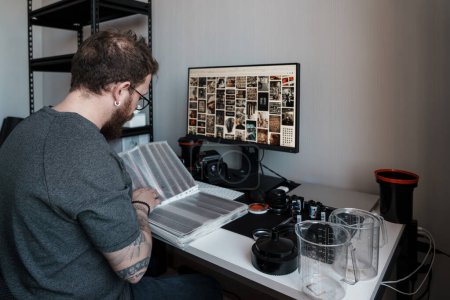 Ein fokussierter Fotograf begutachtet Filmnegative in einem gut ausgestatteten, zeitgenössischen Arbeitsraum voller Fotoausrüstung.