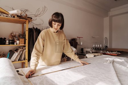 Téléchargez les photos : Une artisan mesure méticuleusement le textile sur une table dans son atelier de couture bien équipé. - en image libre de droit