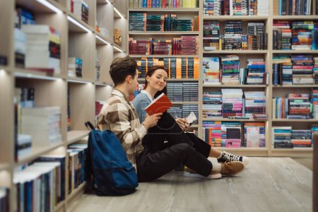 A young man and woman are seated on the floor in a library, surrounded by shelves filled with books. They are smiling and discussing a red book, sharing a peaceful, educational moment.