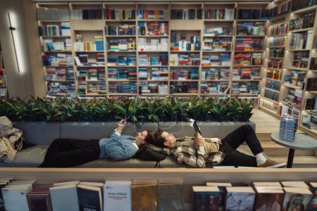 A young couple enjoys a quiet moment, lying down and reading books in a well-stocked modern library surrounded by lush green plants.