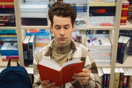 Un jeune homme concentré est profondément absorbé par la lecture d'un livre rouge tout en se tenant dans une librairie entourée d'étagères de titres divers.