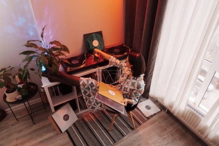 A relaxed young woman listens to vinyl records in a warm, well-decorated interior space.
