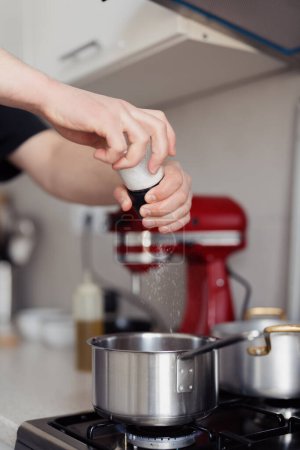 Close-up of hand sprinkling salt into a pot on a stove, in a modern kitchen setting.
