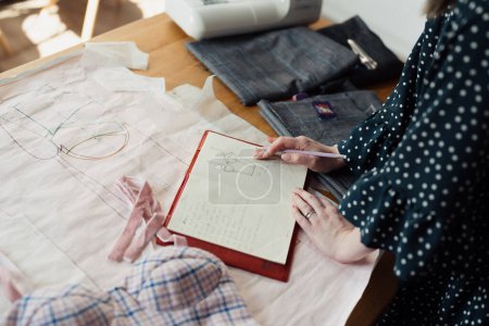 Detail of a fashion designers hands sketching a new garment on pattern paper with fabric samples and a sewing machine in the background.