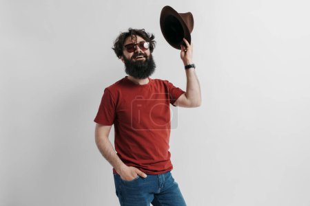 A cheerful bearded man in a red t-shirt and sunglasses is playfully tipping his brown hat. He embodies a casual, relaxed vibe with a hint of hipster style against a neutral background.