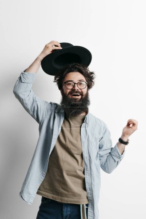 A portrait of a cheerful bearded man in casual clothing tipping his black hat as a friendly gesture, showcasing positive emotions and a welcoming personality against a white background.