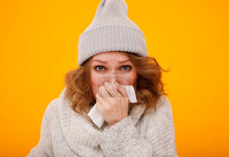 Woman with a cold blowing her runny nose with tissue. Portrait of beautiful girl in winter sweater and hat get sick sneezing from flu. Healthcare and medical concept