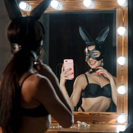 Téléchargez les photos : Femme en masque et oreilles de lapin portant de la lingerie noire assise sur une chaise et faisant du selfie dans le rétroviseur - en image libre de droit