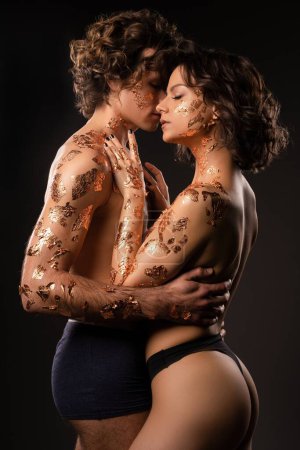 Téléchargez les photos : Vue latérale du couple sensuel topless en feuilles d'or sur les corps embrassant doucement tout en se tenant contre l'obscurité - en image libre de droit