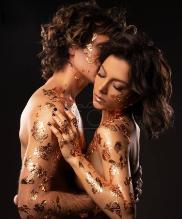 Téléchargez les photos : Vue latérale du couple amoureux sensuel en sous-vêtements aux cheveux foncés ondulés et aux feuilles dorées sur les corps embrassant doucement tout en se tenant debout sur fond noir - en image libre de droit