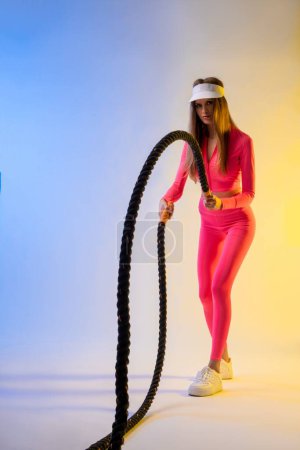 Téléchargez les photos : Corps complet de sérieux jeune modèle féminin avec des cheveux longs dans des vêtements de sport élégants debout dans un studio de couleur vive avec corde de combat - en image libre de droit