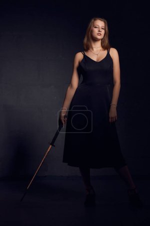 Téléchargez les photos : Corps complet de jeune femme confiante modèle en robe noire avec katana japonais debout dans le studio sombre et regardant la caméra - en image libre de droit