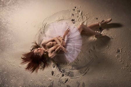 Téléchargez les photos : Vue du dessus de la femme nue sensuelle en jupe de ballet rose et pointe fermer les yeux tout en étant couché sur le sable avec des traces d'ange de sable - en image libre de droit
