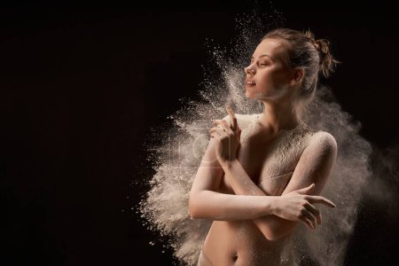 Téléchargez les photos : Jeune femme seins nus touchant les yeux fermés près de la poussière de nuages sur fond noir - en image libre de droit