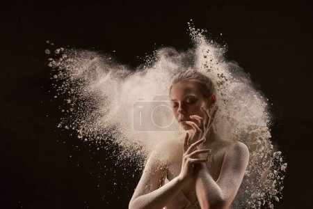 Téléchargez les photos : Jeune femme sereine en sous-vêtements touchant le visage et fermant les yeux près de la poussière de nuage sur fond noir - en image libre de droit