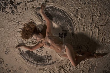 Téléchargez les photos : Vue du dessus de la femme sensuelle en lingerie beige avec les yeux fermés de plaisir tout en étant couché sur le sable avec des traces d'ange de sable - en image libre de droit