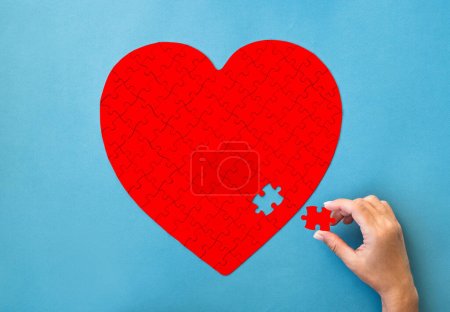 Photo for Red puzzle in heart shape. Hand with white details of puzzle on blue background. - Royalty Free Image