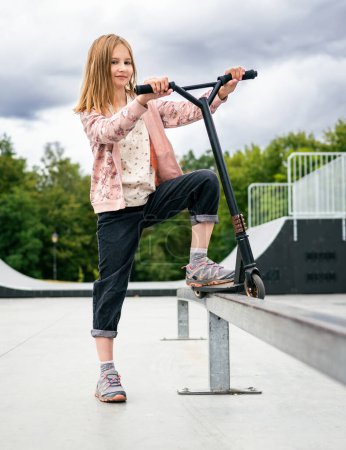 Foto de Chica preadolescente de pie con scooter en la calle en el parque y mirando a la cámara. Niño lindo posando con vehículo ecológico al aire libre - Imagen libre de derechos