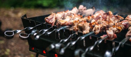 Photo for Barbecue with delicious grilled meat on skewer on brazier outside at summertime. Preparing pork steaks with smoke during picnic at the nature. Closeup view at hot BBQ - Royalty Free Image