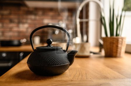 Foto de Black metal teapot with hot tea at the kitchen. Steam comes from the tea spout. Tea drinking traditions. Girl making a tea. - Imagen libre de derechos