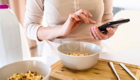 Photo for Girl with oatmeal bowl and smartphone in hands checking social media news during cereal breakfast. Woman with mobile phone and granola at kitchen - Royalty Free Image