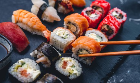 Foto de Set de sushi colorido en escritorio negro con rollo de California. Persona tomar maki con palillo. Cocina oriental japonesa saludable con pescado crudo y arroz - Imagen libre de derechos