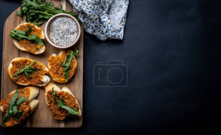 Photo for Mediterranean bruschettas with pesto and arugula served on wooden board with herbal salt and fabric on background with copy space. Traditional italian toasted bread with sauce and plants antipasti - Royalty Free Image