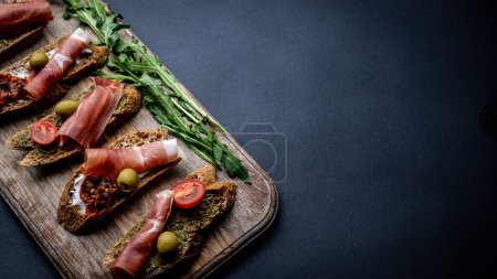 Photo for Bruschettas with jamon, olives, pesto, grilled cherry tomatoes served on wooden board with arugula on background with copy space. Mediterranean toasted bread with meat, cheese and vegetables - Royalty Free Image