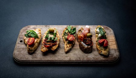 Photo for Set of bruschettas with jamon, olives, pesto, grilled and cherry tomatoes, basil served on wooden board with arugula. Traditional mediterranean toasted bread with cheese, meat and vegetables - Royalty Free Image