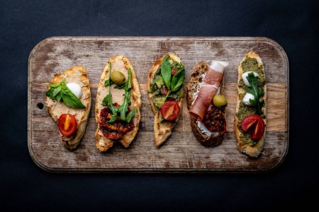 Set of bruschettas with jamon, olives, pesto, grilled and cherry tomatoes, basil served on wooden board with arugula. Traditional mediterranean toasted bread with cheese, meat and vegetables