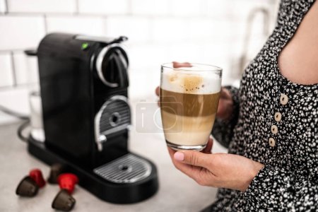 Photo for Girl with cappuccino in transparent cup and capsule coffee machine at kitchen. Woman holding mug with creamy italian caffeine beverage prepared in professional espresso maker - Royalty Free Image