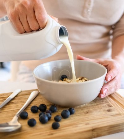 Photo for Girl puts milk to oatmeal with blueberry in bowl plate for delicious cereal breakfast at kitchen. Woman preparing granola with berry and latte for morning healthy diet meal - Royalty Free Image