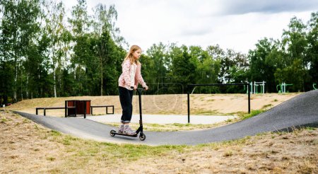 Téléchargez les photos : Preteen fille équitation scooter sur la rampe dans le parc. Enfant mignon avec véhicule écologique à l'extérieur - en image libre de droit