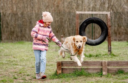 Foto de Chica con perro Golden Retriever en un área de caminata y entrenamiento. Perro saltando sobre la barrera. - Imagen libre de derechos