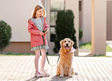 Photo for Girl with golden retriever dog walking outdoors in sunny day in city. Female child kid and pet doggy labrador on lace at street together - Royalty Free Image