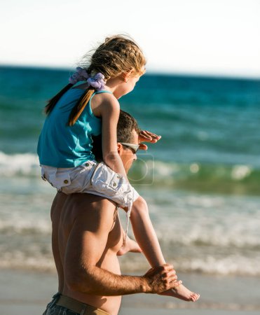 Téléchargez les photos : Père tenant sa fille sur les épaules au bord de la mer et regardant au loin. Papa de la famille et petit joli gosse à la plage à l'été ensemble - en image libre de droit